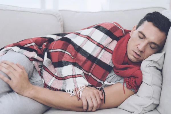 Soğuk bir adam kanepede, kırmızı halı saklanıyor oturuyor. Dijital termometre ile sıcaklık ölçer — Stok fotoğraf