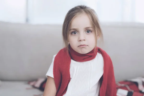 La niña enferma se sienta en un sofá blanco envuelto en una bufanda roja. Tiene una apariencia infeliz. — Foto de Stock