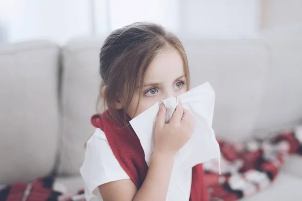 La niña enferma se sienta en un sofá blanco envuelto en una bufanda roja. Se sonó la nariz en una servilleta. —  Fotos de Stock