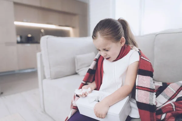 La niña enferma se sienta en un sofá blanco envuelto en una bufanda roja. Estornuda, sacando una servilleta — Foto de Stock