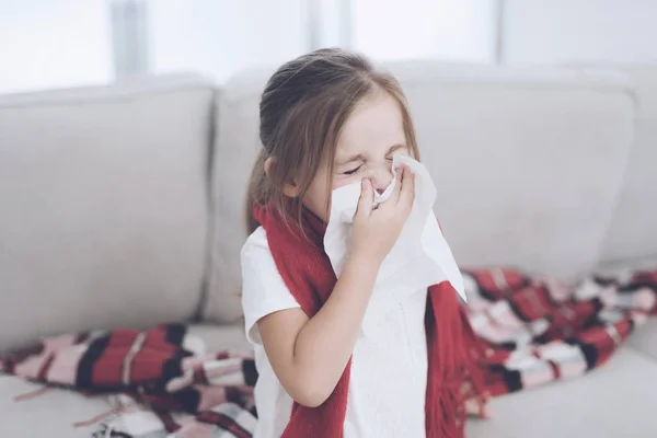 La bambina malata siede su un divano bianco avvolta in una sciarpa rossa. Si soffia il naso in un tovagliolo — Foto Stock