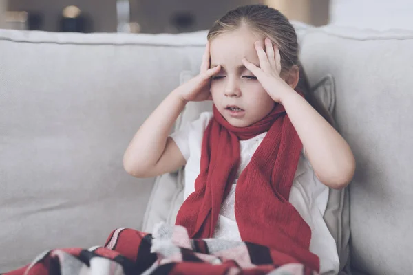 Das kleine kranke Mädchen sitzt auf einem weißen Sofa, das in einen roten Schal gehüllt ist. Sie hat Kopfschmerzen und sehr hohes Fieber — Stockfoto