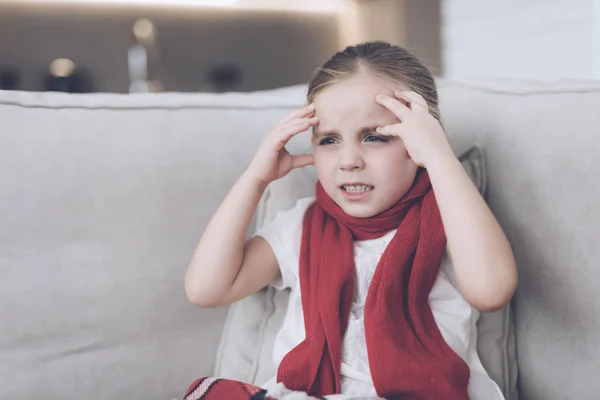 Маленькая больная девочка сидит на белом диване, завернутом в красный шарф. У неё головная боль и очень высокая температура. — стоковое фото