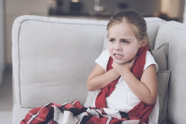 Маленька хвора дівчинка сидить на білому дивані, загорнутому в червоний шарф. У неї дуже болить горло, і вона тримається за нього — стокове фото