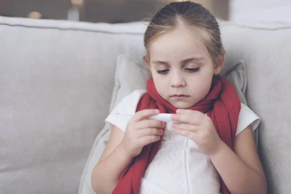 Άρρωστο κοριτσάκι κάθεται σε ένα λευκό καναπέ τυλιγμένο σε ένα κόκκινο μαντήλι. Κοιτάζει το θερμόμετρο, έχει μια υψηλή θερμοκρασία — Φωτογραφία Αρχείου