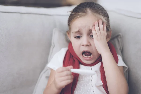 La niña enferma se sienta en un sofá blanco envuelto en una bufanda roja. Mira el termómetro, tiene una alta temperatura — Foto de Stock