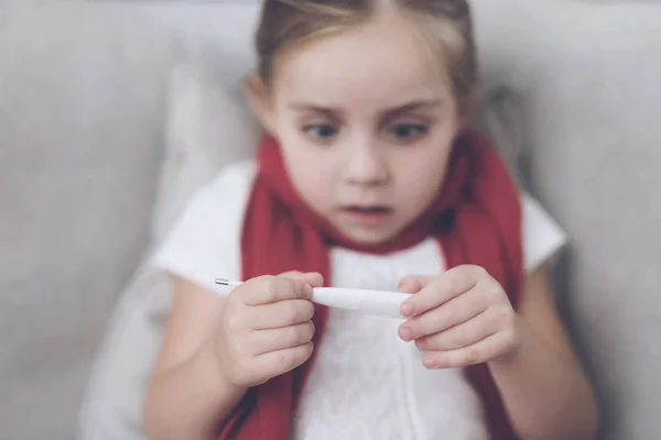 La niña enferma se sienta en un sofá blanco envuelto en una bufanda roja. Mira el termómetro, tiene una alta temperatura — Foto de Stock