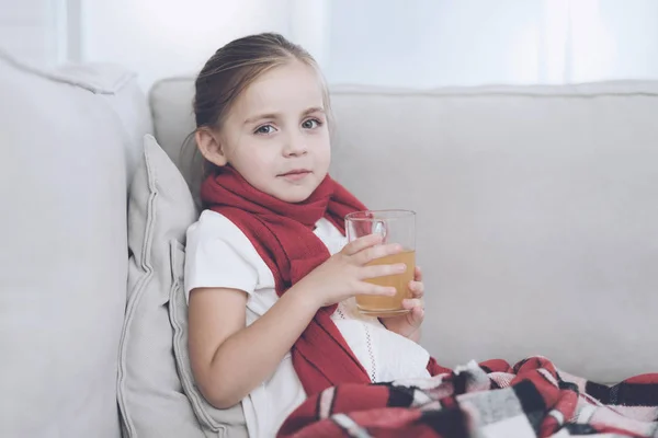 La niña enferma se sienta en un sofá blanco envuelto en una bufanda roja. Ella está sentada con una taza de té medicinal — Foto de Stock