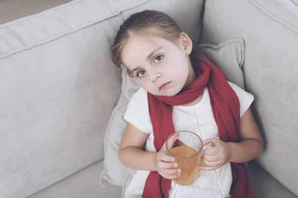 Άρρωστο κοριτσάκι κάθεται σε ένα λευκό καναπέ τυλιγμένο σε ένα κόκκινο μαντήλι. Κάθεται με ένα φλιτζάνι τσάι φαρμακευτικών — Φωτογραφία Αρχείου