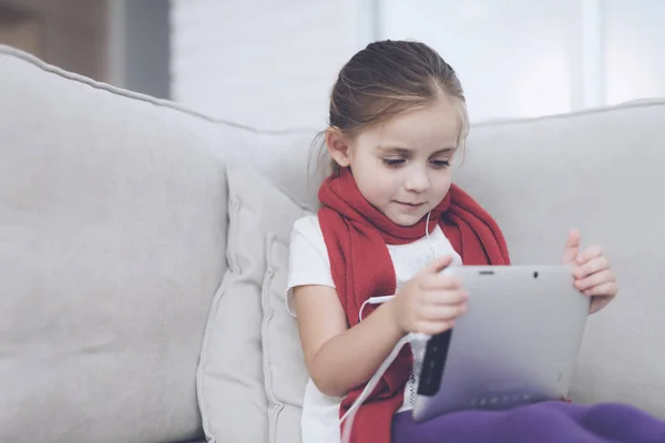 A menina doente senta-se num sofá branco envolto num lenço vermelho. Ela tem um tablet cinzento nas mãos — Fotografia de Stock