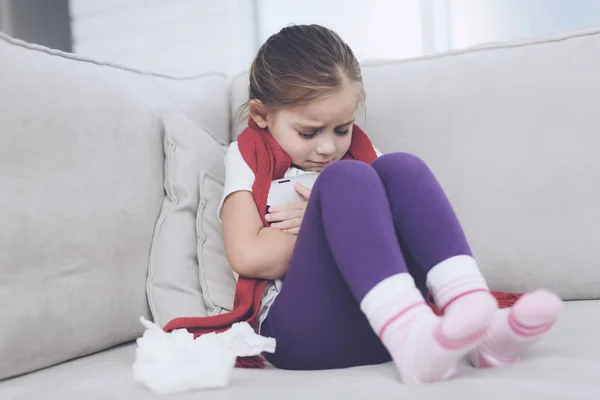 La niña enferma se sienta en un sofá blanco envuelto en una bufanda roja. Ella sostiene una tableta gris en sus manos — Foto de Stock