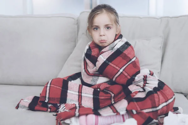 La niña enferma se sienta en un sofá blanco envuelto en una bufanda roja. Tiene frío y se envolvió en una manta roja. — Foto de Stock