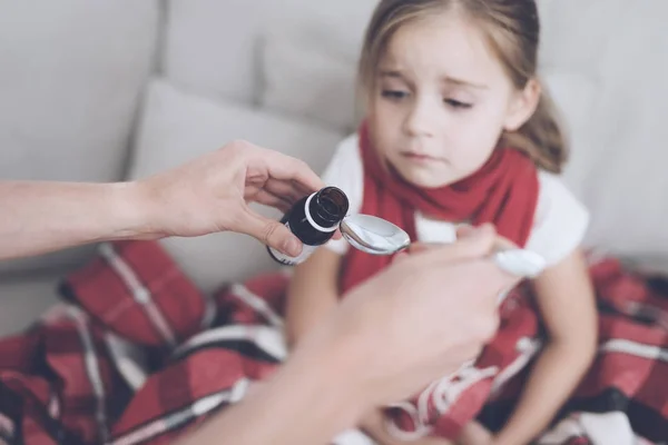 Beteg kislány ül egy fehér kanapén csomagolva egy piros kendő. A szirup ömlik be a kanál — Stock Fotó