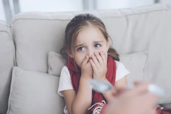 La niña enferma se sienta en un sofá blanco envuelto en una bufanda roja. Ella no quiere tomar el jarabe que se le da — Foto de Stock