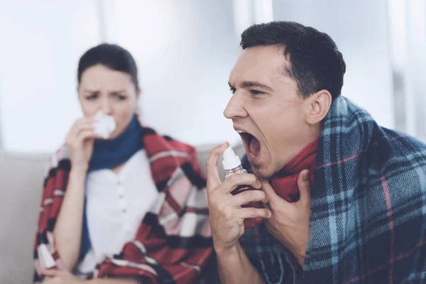 The couple is sitting on the couch wrapped in blankets. The man uses a spray for the throat — Stock Photo, Image