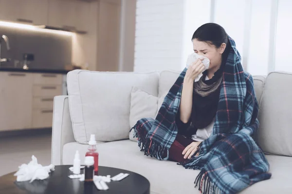 A cold woman sits on a light couch wrapped in a blue checkered whip. She blows her nose into a napkin