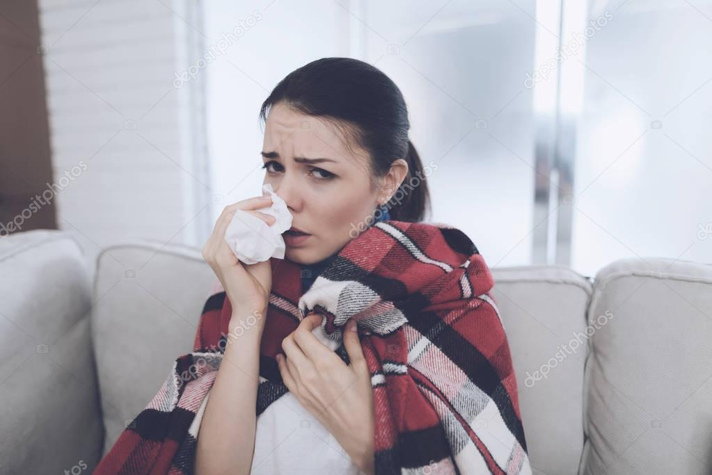 A woman sits on a light couch wrapped in a red plaid plaid. She blows her nose into a paper napkin