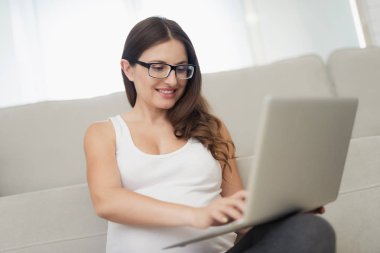 Hamile bir kadın evde hafif bir katta oturuyor. Bacaklarını geçti ve dizüstü bilgisayarda çalışma ile katta oturuyor