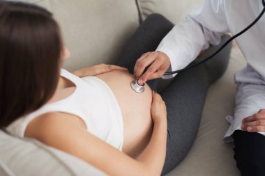 Hamile bir kadın evde ışık kanepede yatıyor. Ona, onunla bir stetoskop ile dinleyen bir doktor geldi