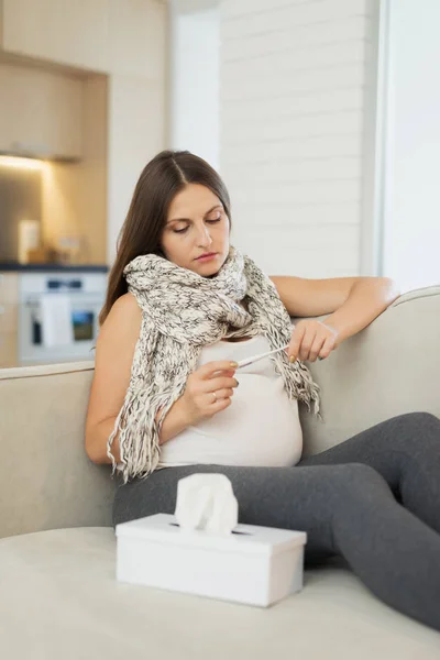 Uma grávida doente sentada em casa no sofá. Ele mede a temperatura com um termômetro eletrônico — Fotografia de Stock