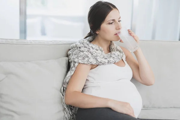 Хвора вагітна жінка сидить вдома на дивані. Вона п'є таблетку, розчинену у воді — стокове фото