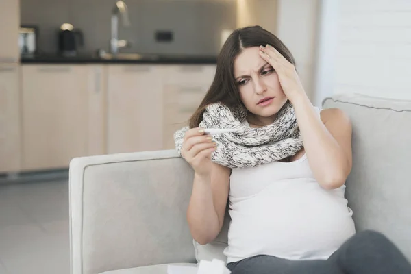 Хвора вагітна жінка сидить вдома на дивані. Вимірює температуру за допомогою електронного термометра — стокове фото