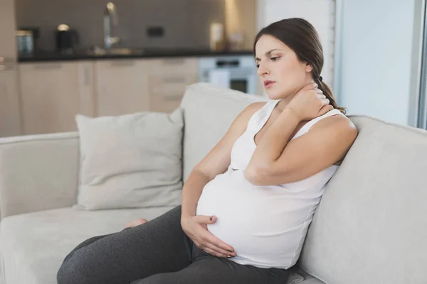 En gravid kvinna sitter hemma på en ljus soffa. Hon har en stel nacke och hon gnuggar det. — Stockfoto