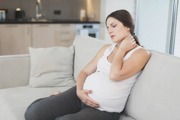 Een zwangere vrouw zit thuis op een lichte sofa. Ze heeft een stijve nek en ze wrijft het. — Stockfoto