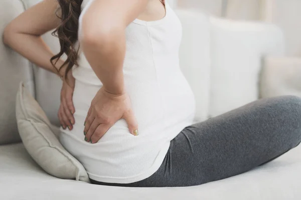 A pregnant woman sits at home on a light sofa. Her back hurts and she holds on to her — Stock Photo, Image