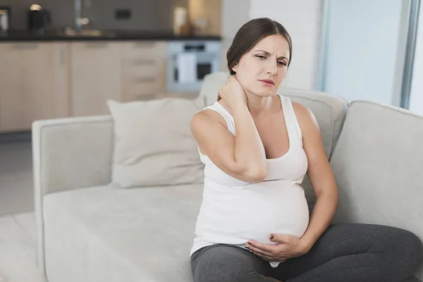 En gravid kvinna sitter hemma på en ljus soffa. Hon håller handen i nacken — Stockfoto