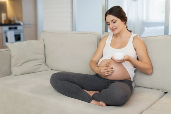 A pregnant woman sits at home on a light sofa. She smears her navel with ointment — Stock Photo, Image