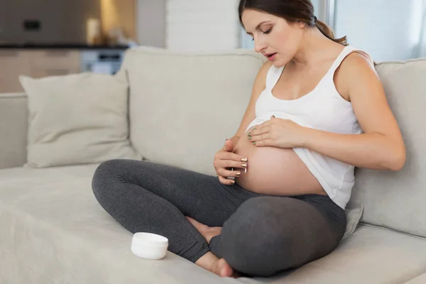 En gravid kvinna sitter hemma på en ljus soffa. Hon smetar sin navel med salva — Stockfoto