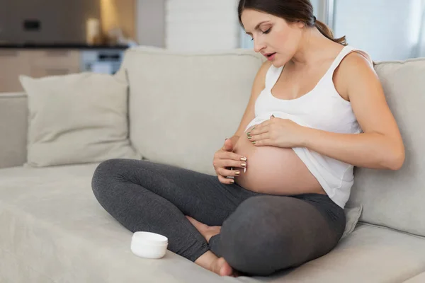 Uma mulher grávida senta-se em casa em um sofá leve. Ela mancha o umbigo com pomada. — Fotografia de Stock