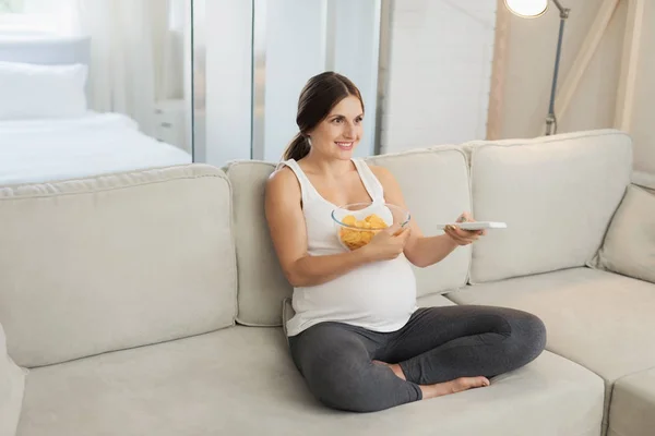 A pregnant woman is sitting on a light sofa at home. She holds a vase of chips and a remote from the TV — Stock Photo, Image