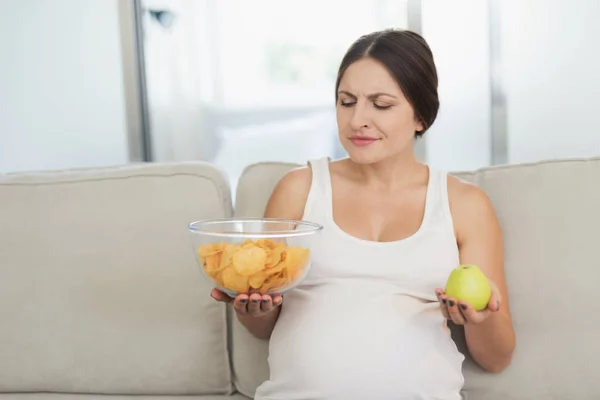 Una mujer embarazada está sentada en un sofá ligero en casa. Ella sostiene un jarrón de papas fritas y una manzana en sus manos —  Fotos de Stock