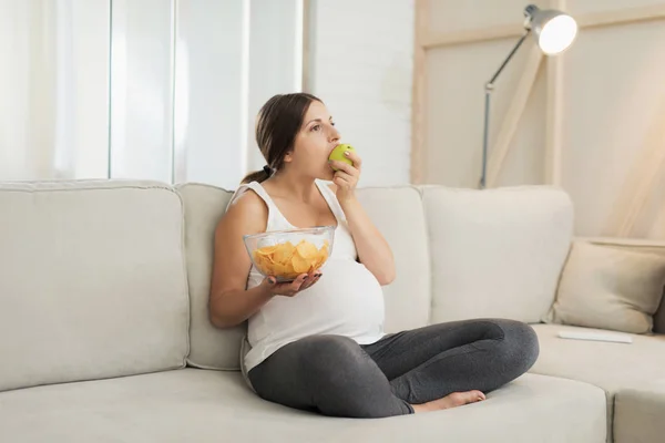Una mujer embarazada está sentada en un sofá ligero en casa. Ella sostiene un jarrón de papas fritas en su mano y se come una manzana — Foto de Stock