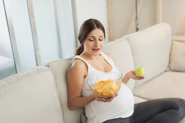 Uma mulher grávida está sentada em um sofá leve em casa. Ela tem um vaso de batatas fritas e uma maçã nas mãos. — Fotografia de Stock