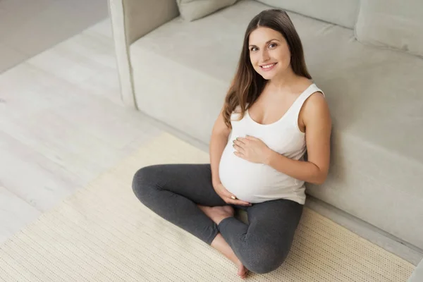 一个孕妇坐在家里的浅色地板上。她坐在地板上, 双腿交叉着, 双手放在肚子上。 — 图库照片
