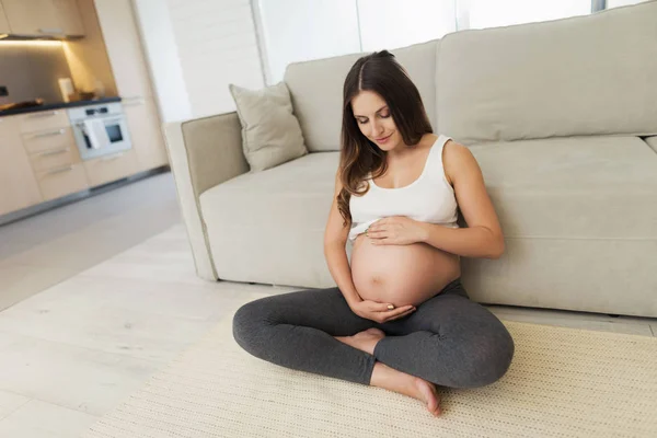 Una donna incinta si siede su un pavimento leggero a casa. Si siede a gambe incrociate sul pavimento e si aggrappa alla pancia — Foto Stock