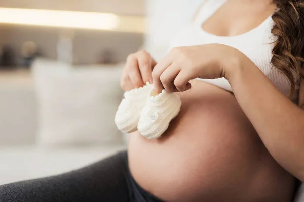 Uma mulher grávida está deitada em um sofá leve em casa. Ela pôs botas de bebé na barriga. — Fotografia de Stock