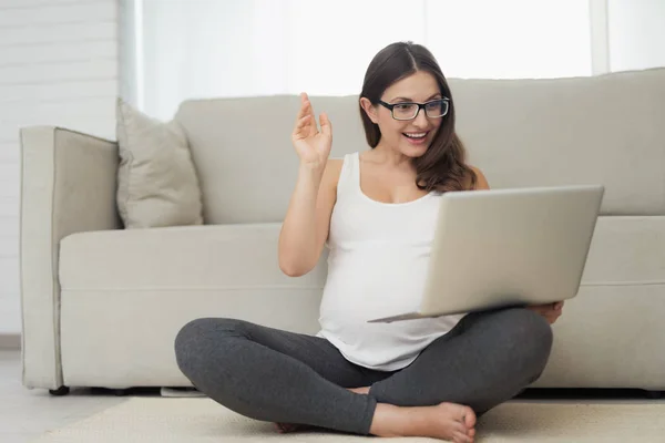 Hamile bir kadın evde hafif bir katta oturuyor. Bacaklarını geçti ve dizüstü bilgisayarda çalışma ile katta oturuyor — Stok fotoğraf
