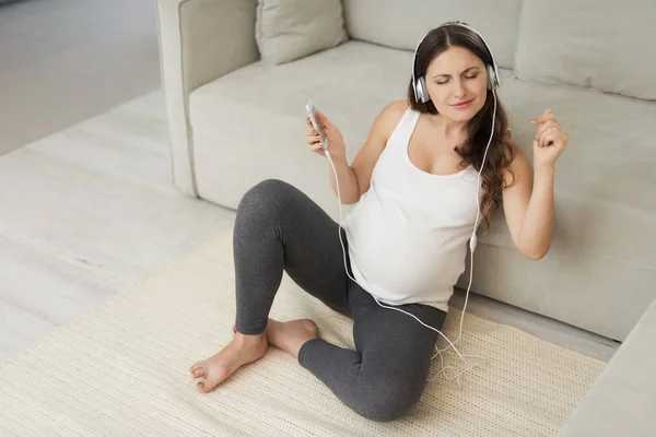 Těhotná žena sedí na světlou podlahou doma. Nosila sluchátka a poslouchat hudbu — Stock fotografie