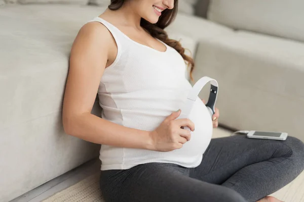 Une femme enceinte est assise sur un sol clair à la maison. Elle a mis les écouteurs à son estomac — Photo