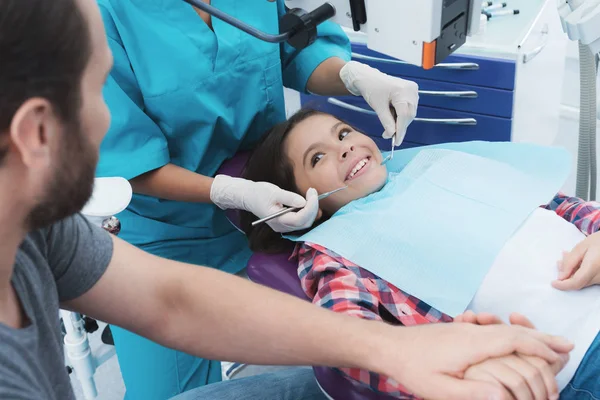 Μια γυναίκα οδοντίατρος μεταχειρίζεται ένα κορίτσι. Κάθεται στην οδοντιατρική καρέκλα. Φοβάται και τον πατέρα της κρατά το χέρι της. — Φωτογραφία Αρχείου