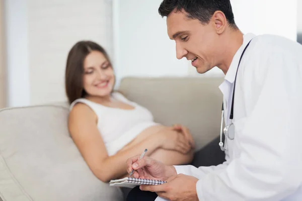 Eine schwangere Frau liegt zu Hause auf einem hellen Sofa. Ein Arzt kam zu ihr, der sie verhörte — Stockfoto