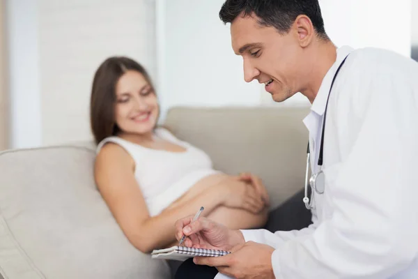 Een zwangere vrouw ligt op een lichte sofa thuis. Een dokter kwam om haar, die haar ondervraagt — Stockfoto