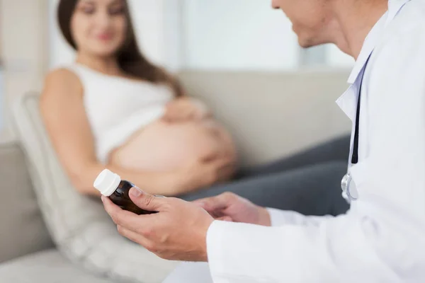 En gravid kvinna ligger i en ljus soffa hemma. En läkare kom till henne, han erbjuder henne någon slags medicinsk produkt — Stockfoto