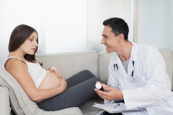 Een zwangere vrouw ligt op een lichte sofa thuis. Een dokter kwam om haar, hij biedt haar een soort medisch product — Stockfoto