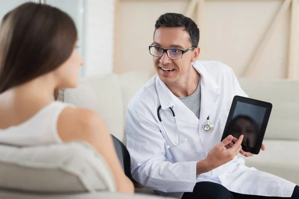 Een zwangere vrouw ligt op een lichte sofa thuis. Een dokter kwam naar haar, hij toont haar iets op haar Tablet PC — Stockfoto