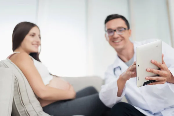 Een zwangere vrouw ligt op een lichte sofa thuis. Een dokter kwam naar haar, hij toont haar iets op haar Tablet PC — Stockfoto